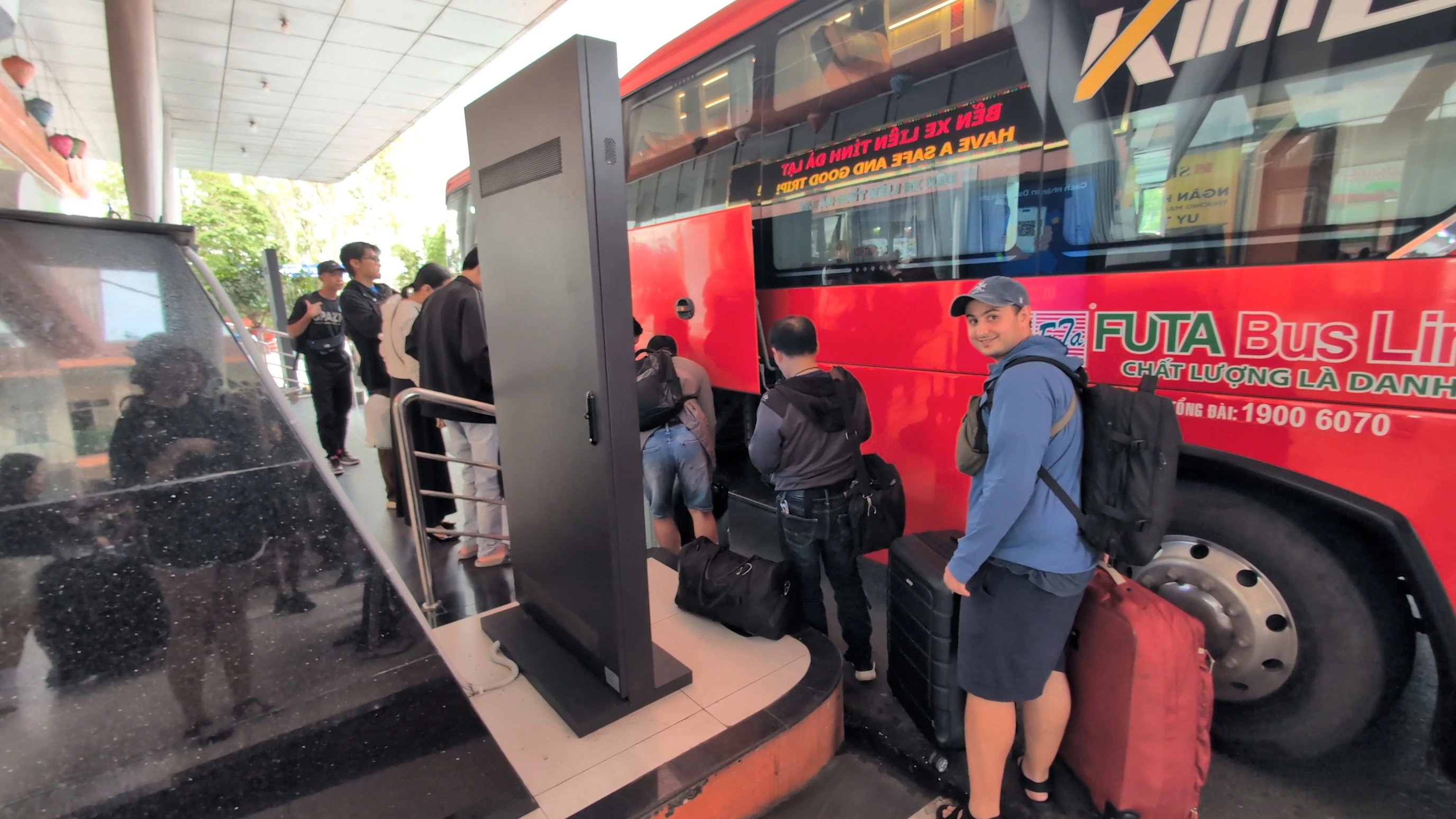 Liam standing with a suitcase beside a red FUTA Bus Lines coach at a busy Vietnam bus terminal