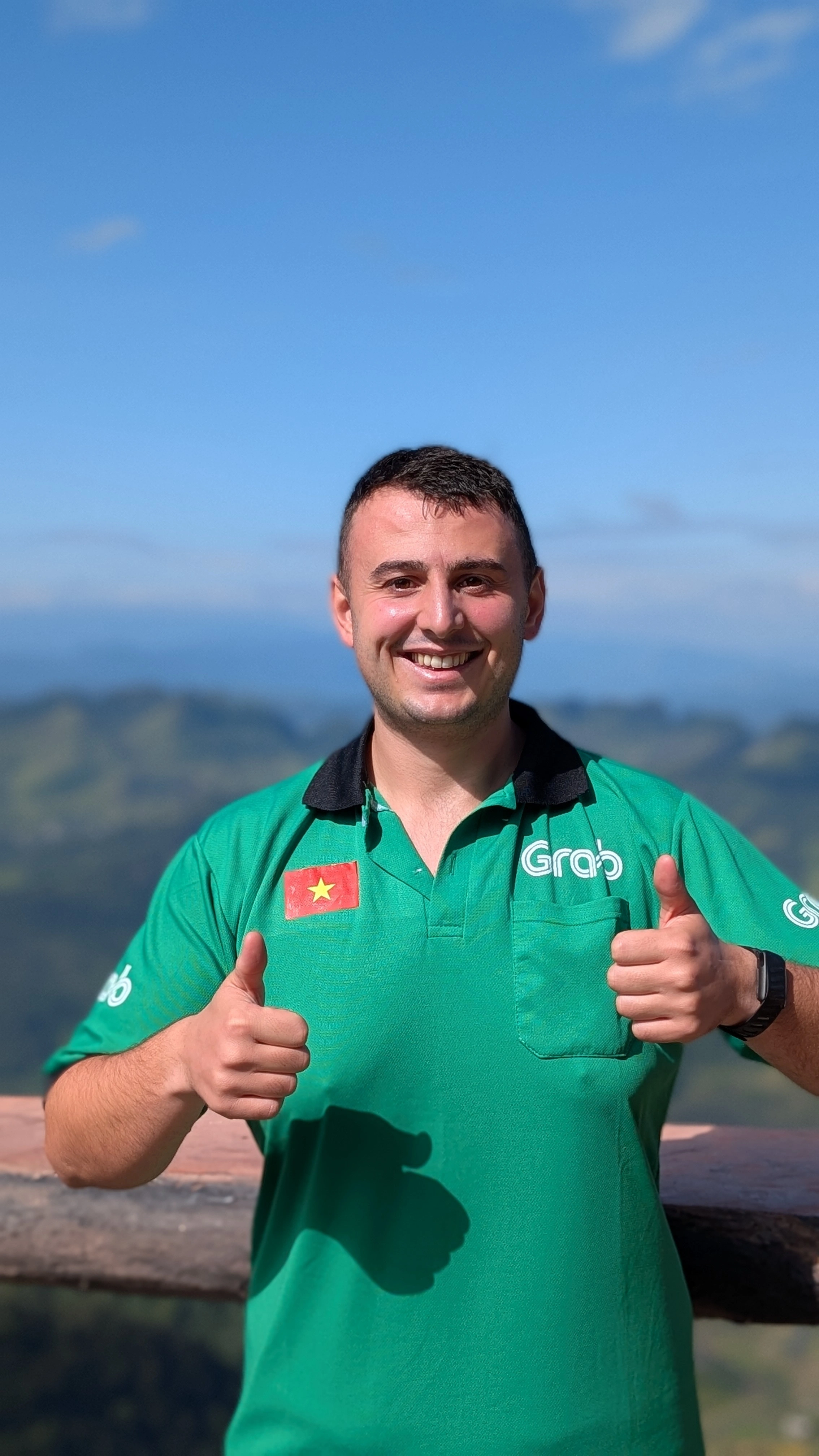 Liam in a green Grab polo shirt giving two thumbs up at a mountain viewpoint in Vietnam