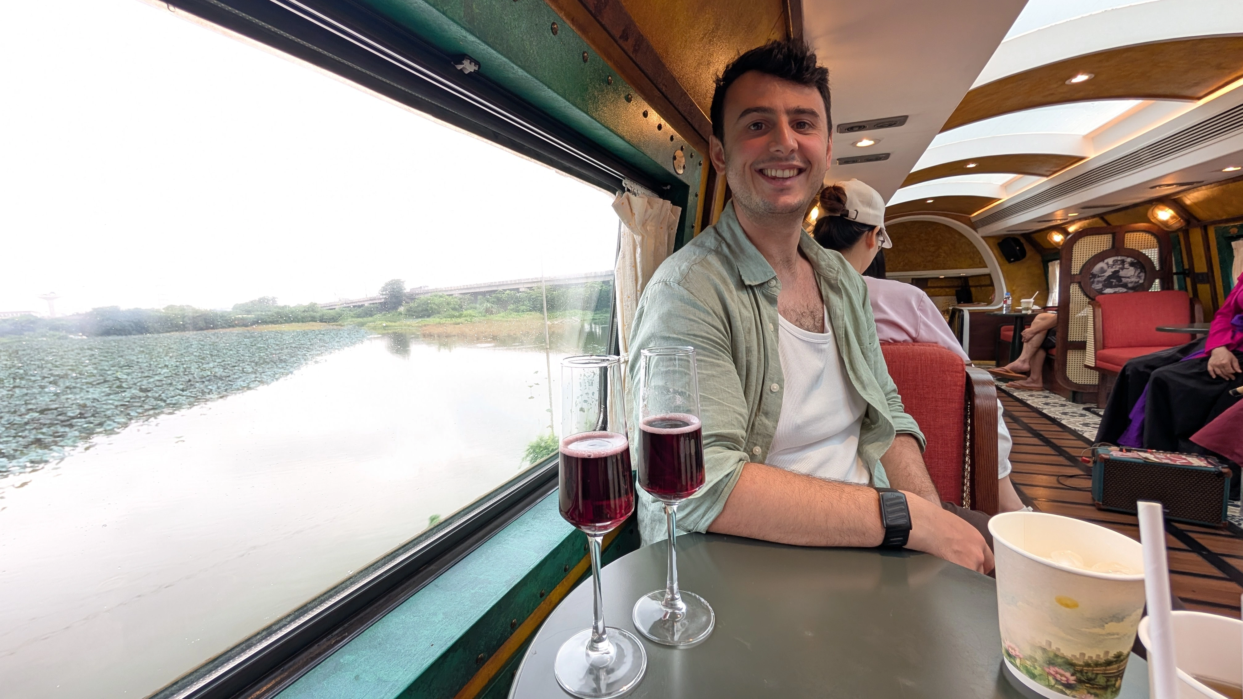 Liam smiling with a drink on a scenic heritage train carriage overlooking green wetlands in Vietnam