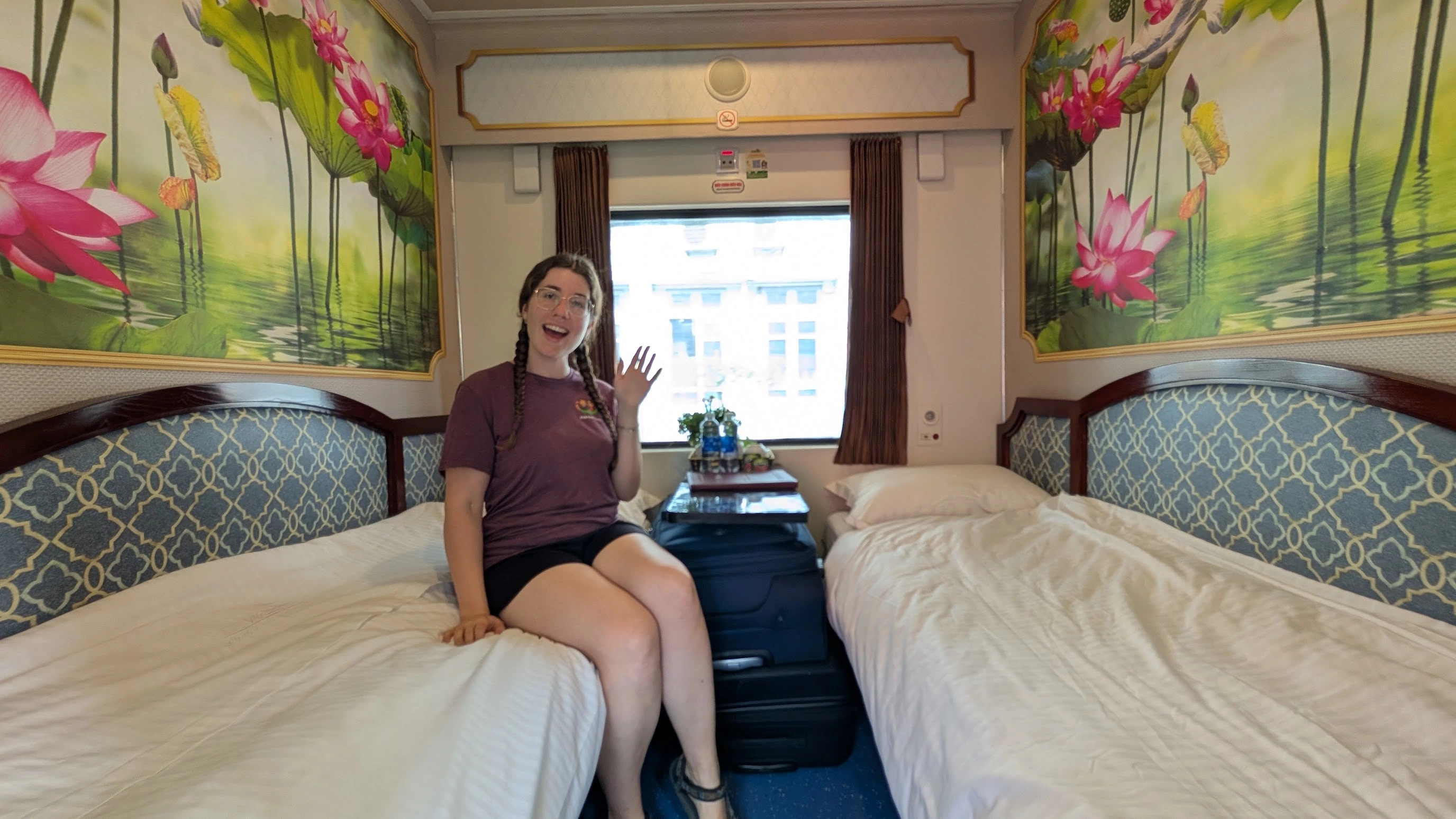 Sarah waving from a clean luxury sleeper train cabin in Vietnam with lotus wall art and twin beds